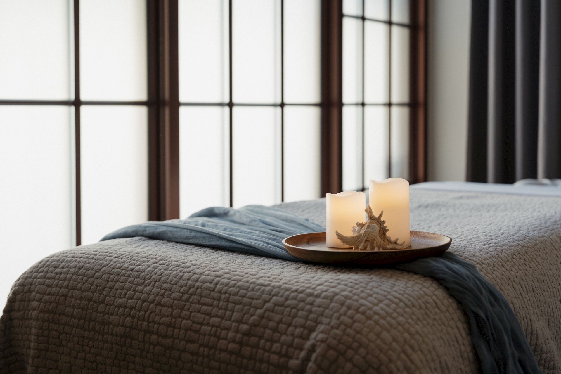 A cozy bed with a folded blanket and two candles on a wooden tray by a window with dark curtains.
