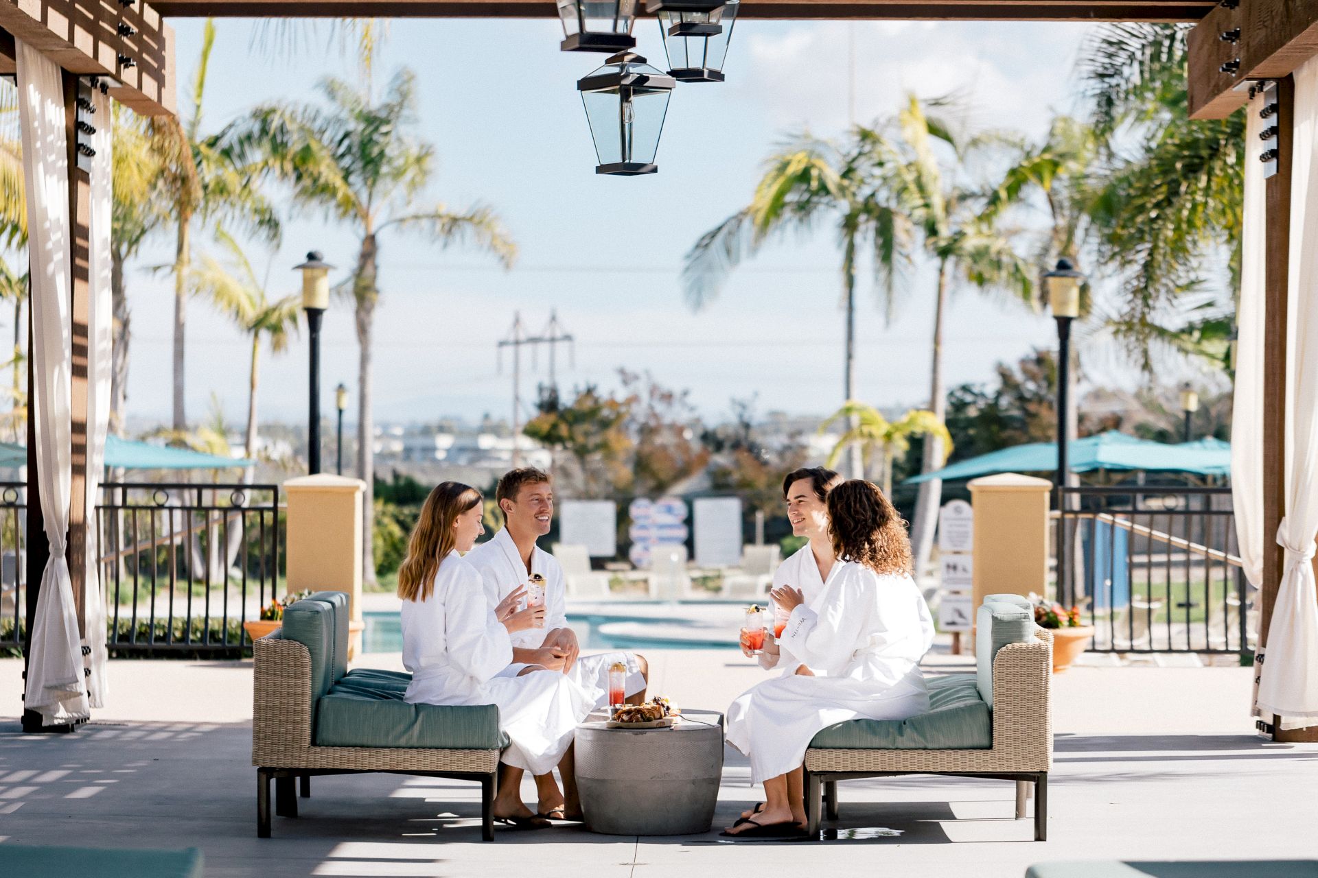 Four friends sit in white robes on a outdoor lounge, chatting and enjoying drinks under a shaded cabana with palm trees and a pool in the background.