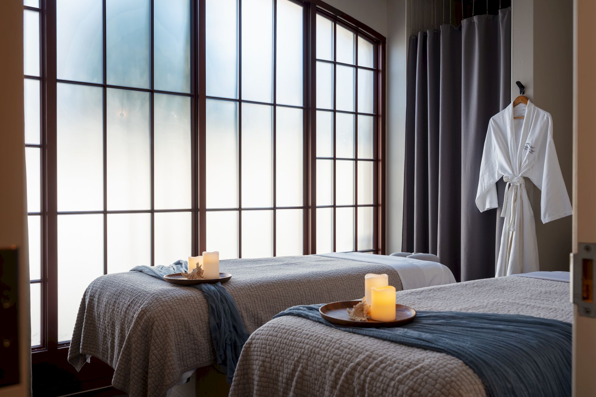 Two spa beds with soft lighting, candles on trays, large grid windows, bathrobes on hanger, serene, tranquil massage room vibes.