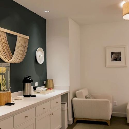 A cozy, modern lounge with a white counter, dark accent wall, a curtain art piece, a coffee station, and white seating in a bright, minimalist space.