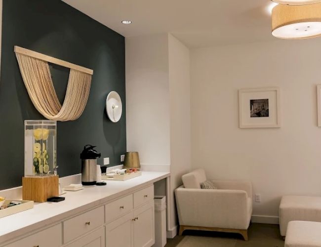 A cozy, modern lounge with a white counter, dark accent wall, a curtain art piece, a coffee station, and white seating in a bright, minimalist space.