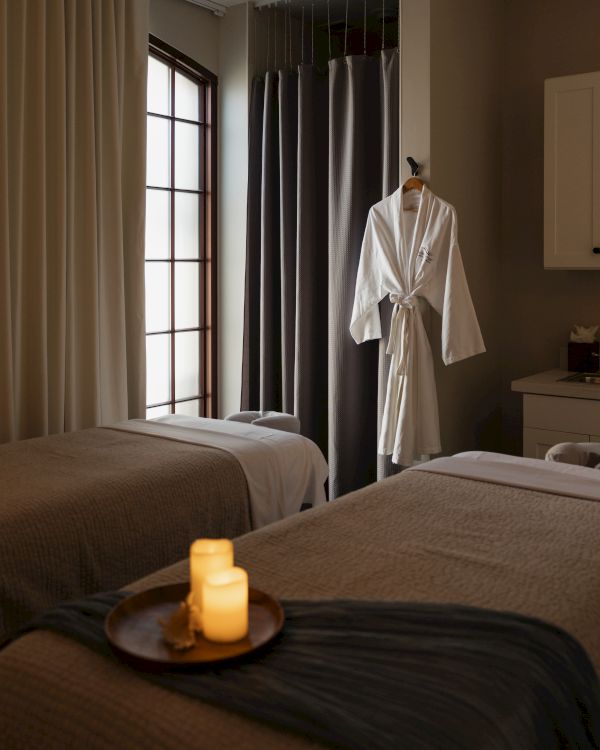 Two neatly made spa-like hotel rooms with soft lighting, candles on a tray, and a white robe hanging by a window and cabinets.