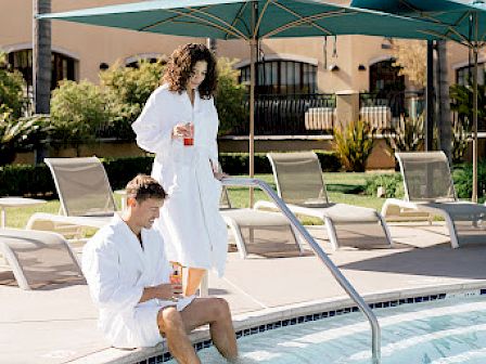 Two people in white robes by a pool, one seated with feet in the water, the other standing with a drink, under sunny skies and palm trees.