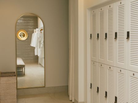 A bright locker room with white lockers on the right, a tall mirror at the end, and a doorway leading to a small seating area. End.