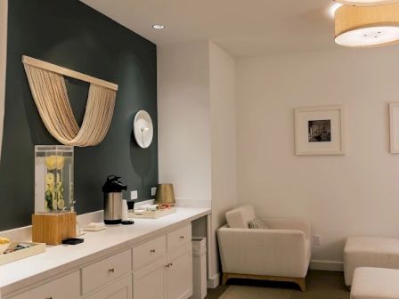 The image shows a cozy room with a white countertop, a water dispenser, coffee pot, seating, wall art, and soft lighting.