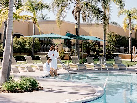 A resort-style pool area with palm trees, lounge chairs, and a couple strolling by the water under blue skies, sunlit and relaxing.